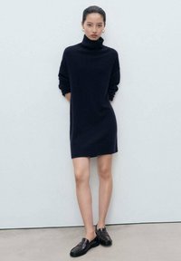 Donna in piedi contro un muro semplice, con indosso un vestito a collo alto nero, mocassini neri, orecchini a cerchio d'oro, con le mani dietro la schiena.