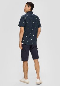 Korte mouwen, donkerblauw shirt met groene sterburstpatronen, gecombineerd met marineblauwe cargoshorts en witte sneakers. Casual ontwerp, lichtgewicht stof.