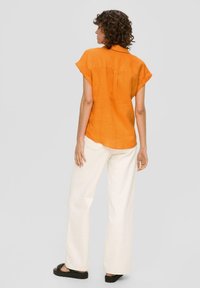 Camicia arancione in lino con colletto e maniche corte, abbinata a pantaloni ampi color avorio e sandali neri. Tessuto morbido, vestibilità casual.