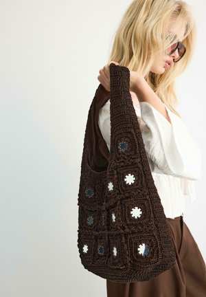Une femme blonde portant des lunettes de soleil tient un sac à bandoulière en crochet marron foncé avec des découpes circulaires sur son épaule.