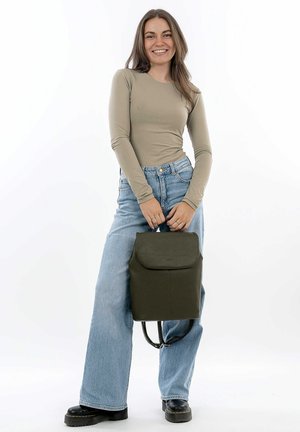 Grüner Leder-Rucksack mit Klappenverschluss, strukturiertem Oberflächenmaterial und verstellbaren Trägern, präsentiert von einer Person in hellen Jeans und einem beigen Top.