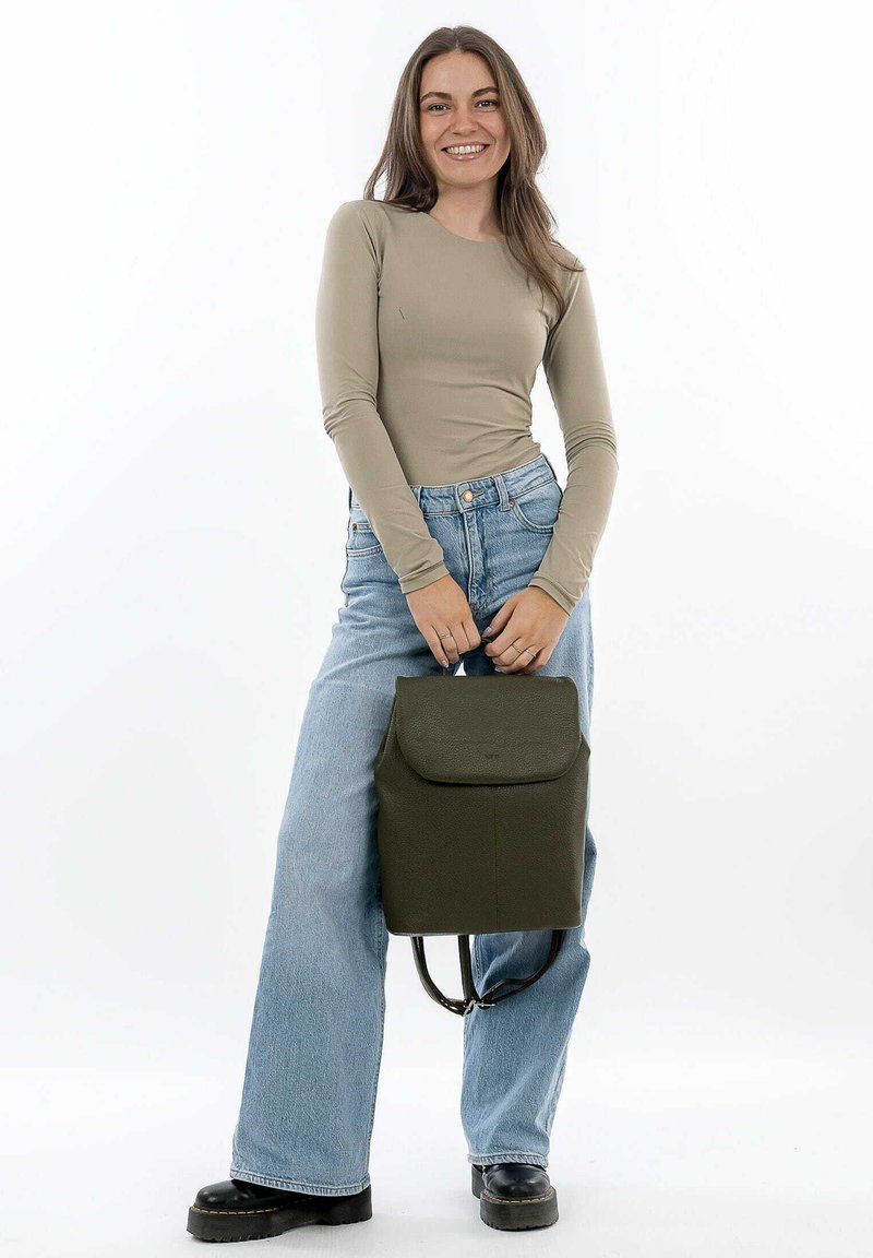 Sac à dos en cuir vert avec un rabat, surface texturée et bretelles réglables, porté par une personne vêtue de jeans clairs et d'un haut beige.