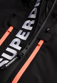 Czarna kurtka z wyraźnym, białym haftowanym logo "SUPERDRY", podwójnymi, jaskrawopomarańczowymi zamkami błyskawicznymi oraz teksturowanym wykończeniem materiału.