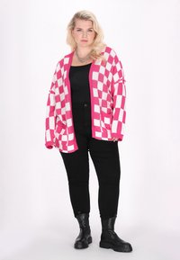 Rosa och vit rutmönstrad stickad cardigan med neddragna ärmar och fickor, kombinerad med svarta åtsittande byxor och kraftiga svarta kängor.