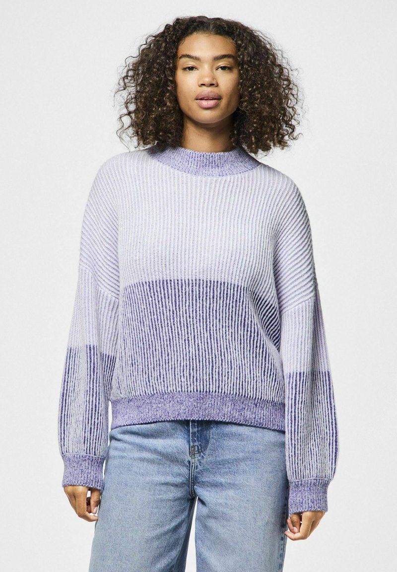 Ljubičasti džemper s prugama, visokog ovratnika, rebraste teksture, dugih rukava i opuštenog kroja, uparen sa svijetloplavim trapericama.