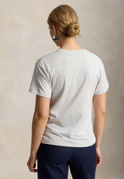 Žena se světlými vlasy svázanými do drdolu, oblečená v světle šedém tričku s krátkým rukávem a tmavých kalhotách, čelem odvrácená od jednoduchého pozadí.