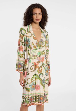 Vrouw met krullend donker haar, gekleed in een knie-lange crème jurk met lange mouwen en kleurrijke bloem- en rankpatronen.
