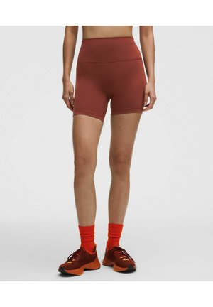 Personne portant un short ajusté taille haute couleur rouille, des chaussettes orange vif et des chaussures de sport rouge foncé, debout sur un fond clair.
