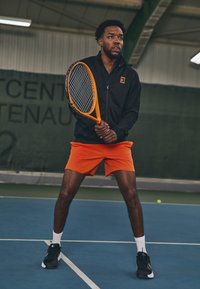 Tenista v čiernej bundě na zips s logom, oranžových šortkách, čiernych teniskách a bielych ponožkách na modrom kurte, ktorý drží oranžový tenisový raketu.