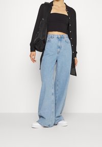 Donna che indossa jeans a gamba larga blu chiaro, top nero senza spalline, camicia aperta nera, sneakers bianche e porta una borsa a tracolla nera.