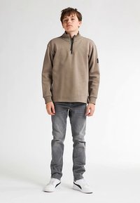 Hanorac bej deschis cu fermoar și guler înalt, purtat peste blugi gri. Are o etichetă neagră pe mâneca stângă și adidași albi.