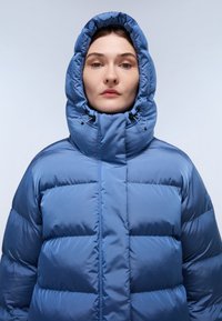Giacca a vento blu con cappuccio alto regolabile. Realizzata in materiale liscio e lucido con texture trapuntata e pannelli orizzontali.