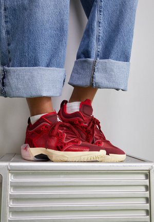 Personne portant un jean bleu retroussé et des chaussettes blanches avec des baskets Jordan rouges et beiges, debout sur une surface métallique.
