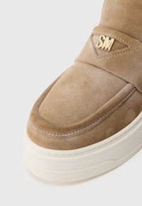 Pantof bej din suede cu o textură netedă, având un logo auriu „SM” proeminent și o talpă din cauciuc albă. Detalii de cusătură vizibile în jurul marginilor.