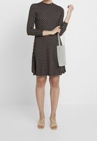 Kvinna i svart mönstrad långärmad klänning som håller en vit pärlhandske, bärande gula snörade högklackade sandaler, stående mot en vit bakgrund.