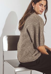 Adīts pārspīlēts džemperis brūnās krāsās ar teksturētu apdari, ar īsām piedurknēm. Modelis sēž uz metāla krēsla, rādīdams sānu profilu.