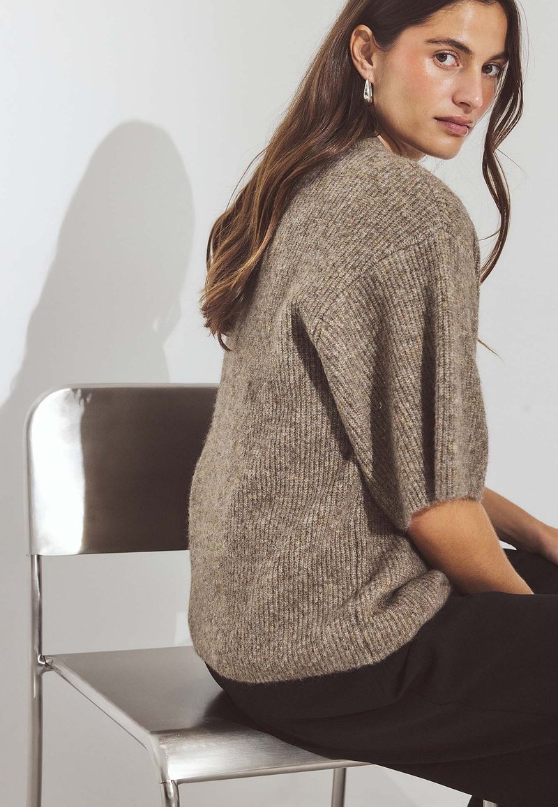 Adīts pārspīlēts džemperis brūnās krāsās ar teksturētu apdari, ar īsām piedurknēm. Modelis sēž uz metāla krēsla, rādīdams sānu profilu.