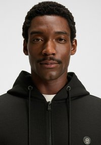 Primo piano di un uomo con capelli ricci corti e baffi, che indossa una felpa nera con zip e un logo ricamato sul petto.