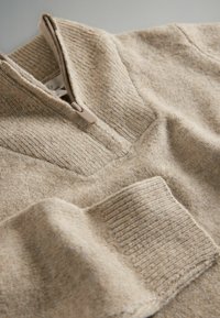 Beżowy sweter z ćwierć zamkiem, wykonany z miękkiego dzianinowego materiału. Posiada ściągacze oraz fakturowe akcenty materiałowe.