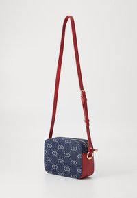 Crossbody-Tasche mit einem marineblauen Denim-Außenmaterial, das ein sich wiederholendes Logo-Muster aufweist, einem roten Lederriemen, Reißverschluss und goldfarbenen Beschlägen.