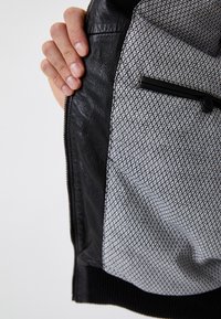 Czarna skórzana kurtka z wzorzystym szarym wnętrzem, z zamkiem błyskawicznym i kieszenią z czarnym guzikiem. Teksturowane wykończenie, elegancki design.
