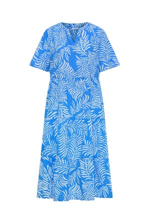 Femme aux cheveux courts et bruns portant une robe bleue mi-longue à manches courtes avec des motifs de feuilles blanches, debout devant un fond beige uni.