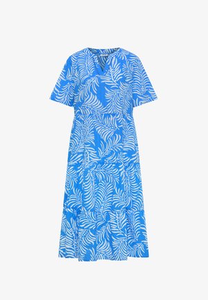 Femme aux cheveux courts et bruns portant une robe bleue mi-longue à manches courtes avec des motifs de feuilles blanches, debout devant un fond beige uni.