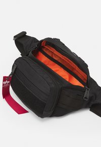 Fanny pack de tecido preto com um interior laranja brilhante, um bolso frontal com fecho e fita ajustável. Apresenta velcro e uma etiqueta.