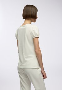 T-shirt color crema con maniche a sbuffo corte, scollatura rotonda e una texture morbida. Indossato con pantaloni chiari coordinati.