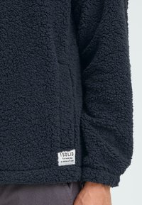 Pullover in pile blu navy con una consistenza soffice, dotato di un piccolo etichetta bianca in basso a sinistra con nome del marchio e slogan.