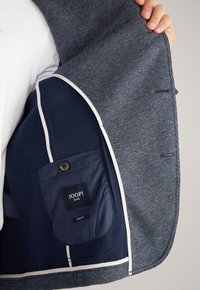 Navy blue blazer with a textured fabric. Features a contrasting inner pocket with a button and a label. Slim fit design with white piping.