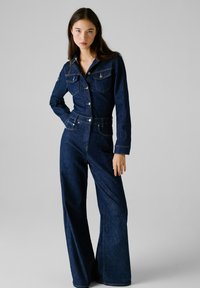 Combinaison en denim à manches longues, avec un devant boutonné, deux poches poitrine et une silhouette à jambes larges de couleur bleu foncé. Elle possède une taille ceinturée.
