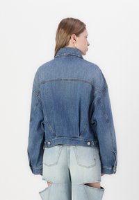 Veste en denim bleu avec une coupe décontractée, grand col et détails de couture visibles. Dotée de boutons-pression et de coutures contrastantes.