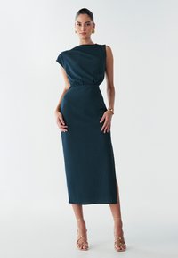Robe en vert d'eau, de longueur genoux, avec une épaule drapée et une taille élastique. Fabriquée en tissu texturé, avec une fente latérale et associée à des talons à lanières.