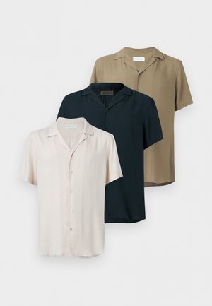 Homme portant une chemise beige à manches courtes boutonnée, un pantalon blanc, des chaussures marron sans lacets et des lunettes de soleil, debout avec une main dans la poche.