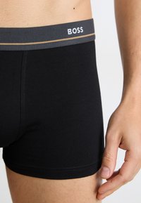 Boxer shorts en coton noir avec une taille gris affichant le logo "BOSS" en blanc et une fine bande d'accent doré. Texture lisse.