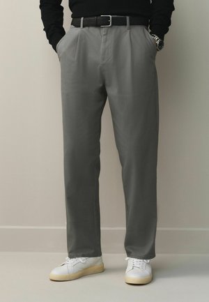 Mann trägt graue plissierte Hose mit schwarzem Gürtel, schwarzes Langarmoberteil, weiße Turnschuhe und eine Armbanduhr, Hände in den Taschen, stehend auf hellem Boden.