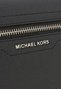 Juodas odinis piniginė su tekstūruotu paviršiumi, turinti sidabrinę užtrauktuką, siuvinėjimo detales ir iškilų "MICHAEL KORS" logotipą.