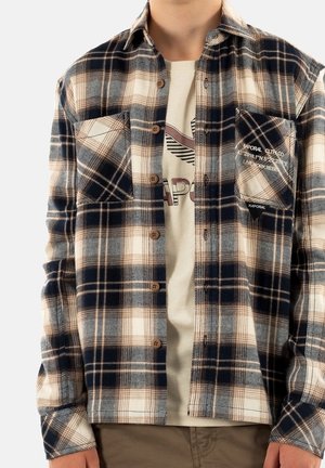 Osoba nosząca beżową i granatową kraciastą koszulę na guziki na jasnobeżowym t-shircie z nadrukiem, u dołu widoczne spodnie khaki.