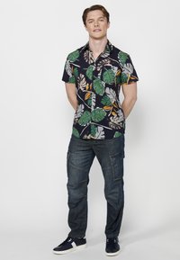 Camisa de manga corta azul marino con un patrón de hojas verde y beige, combinada con pantalones cargo azules y zapatillas azul marino con acentos blancos.