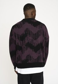 Lila und schwarzer Pullover mit einem Zickzackmuster, dickem Stricktextil und gerippten Bündchen. Über einem weißen Hemd getragen.