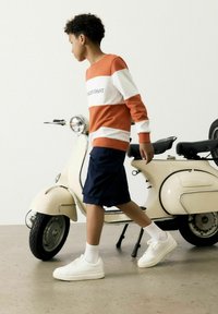 Sweatshirt rayé orange et blanc avec le texte "CALIFCOAST", shorts navy, baskets blanches et une vieille trottinette crème en arrière-plan.