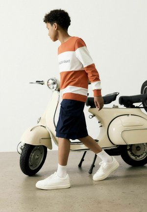 Oranje en wit gestreepte sweatshirt met de tekst "CALIFCOAST", marineblauwe shorts, witte sneakers en een vintage crèmekleurige scooter op de achtergrond.