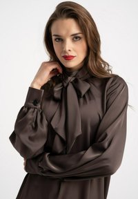 Brązowa jedwabna bluzka z kołnierzykiem wiązanym na kokardę, bufkami na długich rękawach i zapinanymi mankietami. Gładka faktura i wyraźny detal układającego się materiału.