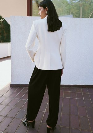 Mujer de cabello oscuro con blazer blanco, pantalones negros de pierna ancha y tacones negros con lazos, de pie sobre un suelo de azulejos cerca de una pared blanca al aire libre.