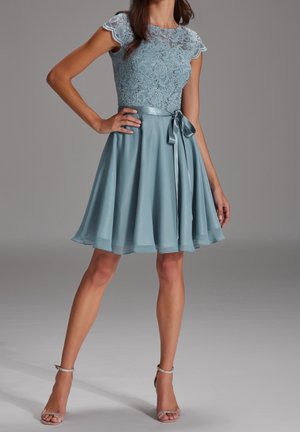 Kvinde iført en knælange, lyseblå blondekjole med korte ærmer og bælte af satinbånd, kombineret med sølvfarvede sko med ankelrem.