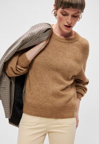 Brauner Strickpullover mit rundem Ausschnitt und geripptem Saum. Getragen mit cremefarbenen Hose und einer karierter Jacke über der Schulter.