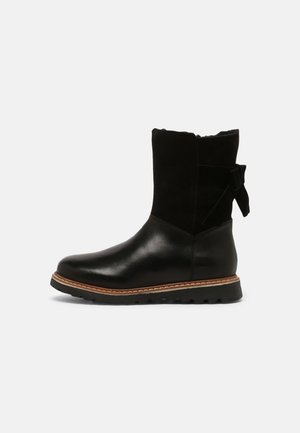 Botte noire mi-mollet avec partie inférieure en cuir lisse, partie supérieure en daim, détail d'un nœud sur le côté, bordure marron au-dessus de la semelle robuste et bout arrondi.