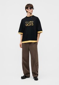 Junger Mann, der ein schwarzes, übergroßes T-Shirt mit gelben Kanten und Leopardenmuster-Text, braune Cargohosen, schwarze Schuhe und übergroße Sonnenbrillen trägt.