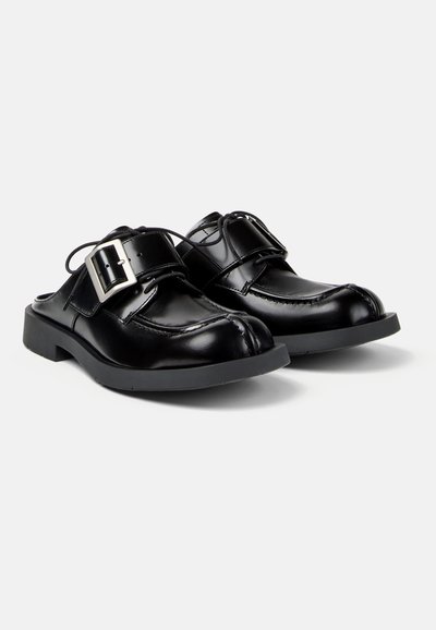 Chaussures en cuir noir à enfiler avec de grandes boucles argentées, bouts carrés arrondis et semelles en caoutchouc épaisses sur un fond uni.